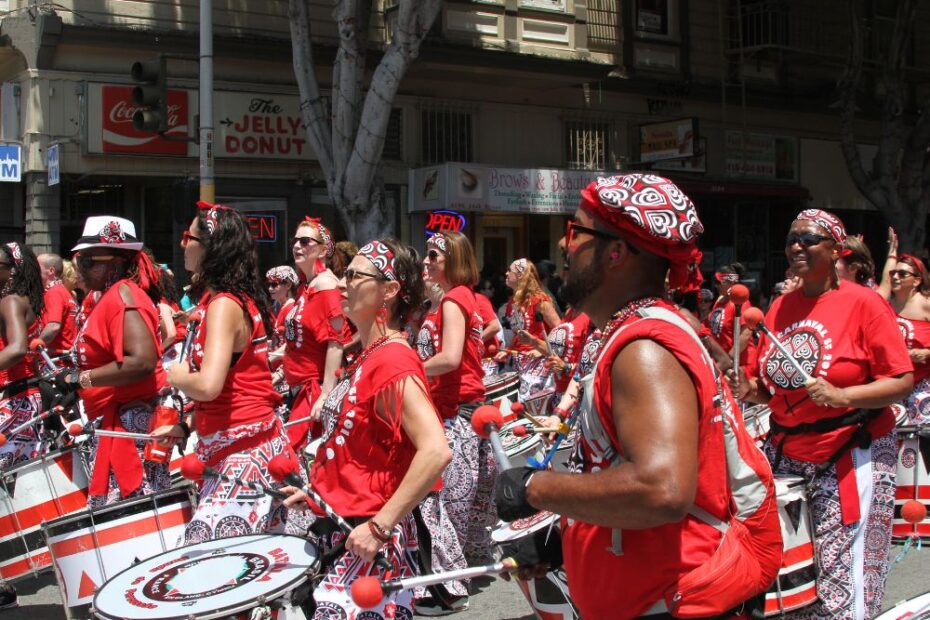 Grupo de percussionistas fantasiados com roupas vermelhas e estampadas toca tambores durante desfile de carnaval em uma grande cidade.